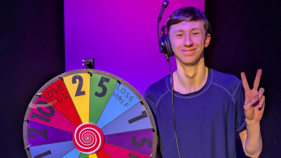 Josh holding a peace sign next to a multi coloured gameshow wheel.