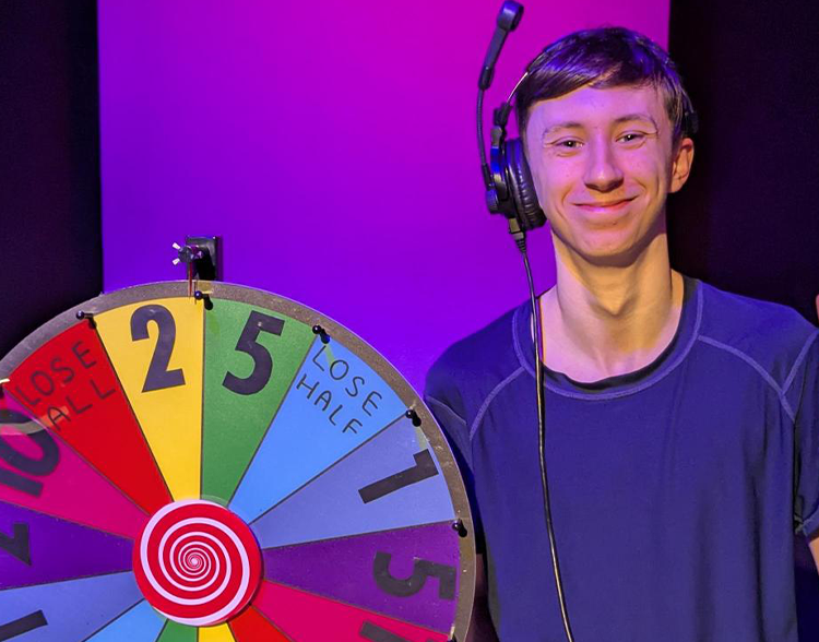Josh holding a peace sign next to a multi coloured gameshow wheel.