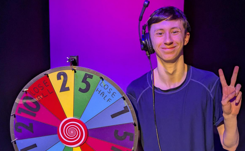 Josh holding a peace sign next to a multi coloured gameshow wheel.