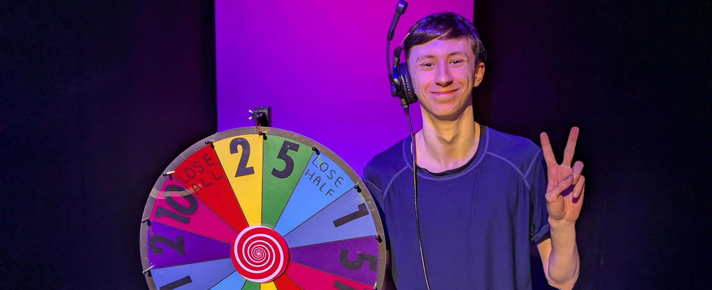 Josh holding a peace sign next to a multi coloured gameshow wheel.