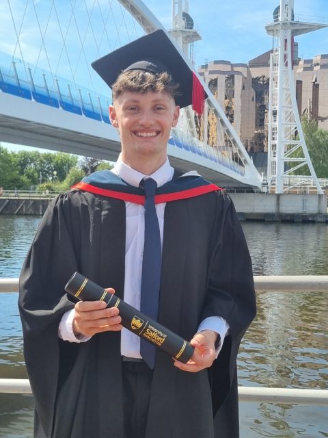 Joshua Saxon in graduation robes with scroll box outside, smiling